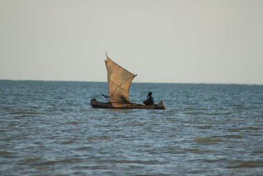 Ein einzelner Fischer sitzt in einem traditionellen Boot mit einem großen, durchsichtigen Segel auf dem ruhigen Wasser.
