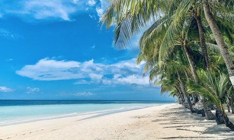 Ein ruhiger Strand mit feinem, weißen Sand erstreckt sich unter einem strahlend blauen Himmel und sanften Wolken.