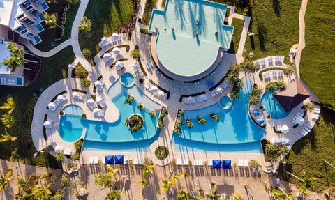 Ein atemberaubender Blick auf einen Poolbereich mit mehreren Schwimmbecken und Liegen, umgeben von Palmen und grünem Gras.
