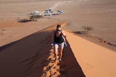 Eine Mutter läuft mit ihrem Kind auf dem Rücken eine Düne hinauf - Namibia Reise mit Kindern