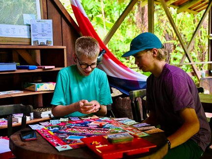 Zwei Jugendliche sitzen an einem Tisch und spielen Monopoly, umgeben von einer einladenden Umgebung mit grünem Pflanzenbewuchs.