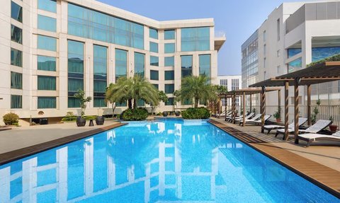 Ein glitzernder Pool spiegelt die moderne Architektur des Pride Plaza Hotels in Neu-Delhi wider, umgeben von Palmen.