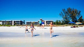 Drei Kinder laufen fröhlich am Strand entlang, während ihre Familie im Hintergrund entspannt. Strahlend blauer Himmel über ihnen.