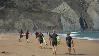 Eine Gruppe von Surfern in Neoprenanzügen geht am Strand entlang, während die Wellen sanft ans Ufer rollen.
