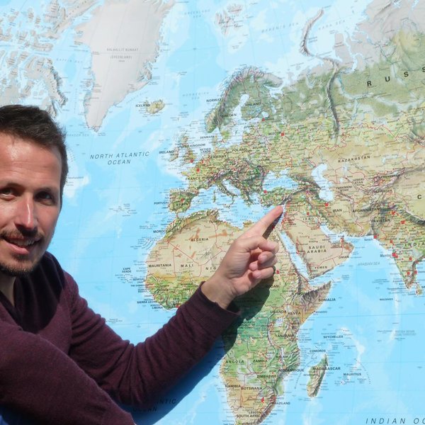 Ein Mann sitzt vor einer Weltkarte und zeigt mit dem Finger auf einen bestimmten Ort in Europa.