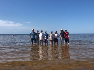 Eine Gruppe von acht Personen steht im flachen Wasser, lächelnd, mit dem Meer im Hintergrund und strahlendem Sonnenschein.