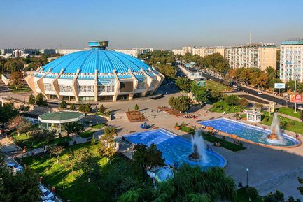 Ein großer, blauer Zirkus mit einer markanten Kuppel steht in einer lebhaften Stadtlandschaft mit Bäumen und Gebäuden.