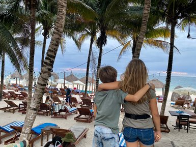 Zwei Kinder stehen am Strand, umarmt, während sie auf das Meer und die Liegen blicken, umgeben von Palmen.
