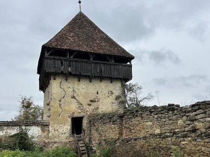 Ein alter, verfallener Turm mit einem schrägen Dach steht inmitten von hohem Gras und umgeben von einer steinernen Mauer.