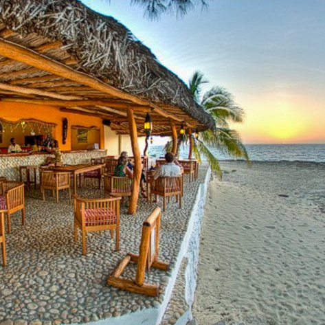 Ein einladendes Strandrestaurant mit Holzstühlen und einem Blick auf den Sonnenuntergang über dem Meer.