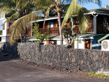 Ein Holzhaus mit mehreren Balkonen, umgeben von Palmen und einem Steinmauer, steht in einer ruhigen Umgebung.