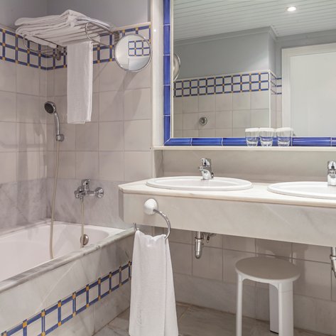 Ein modernes Badezimmer mit einer Badewanne, zwei Waschbecken und einem großen Spiegel, umgeben von hellen Fliesen.