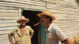 Zwei ältere Männer stehen vor einem Holzhaus, einer trägt einen Strohhut und hat eine Zigarre im Mund.