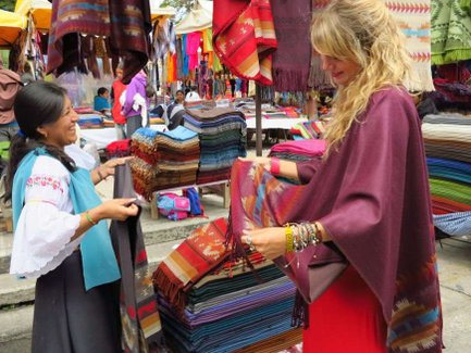 Eine lächelnde Frau in traditioneller Kleidung zeigt einem Touristen bunte Textilien auf einem Markt in Otavalo.