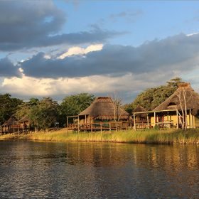Eine friedliche Landschaft mit traditionellen Hütten am Ufer eines ruhigen Gewässers, umgeben von üppigem Grün.