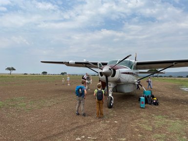 Ein kleines Flugzeug steht auf einem offenen Feld, während Kinder und Erwachsene sich um das Gepäck versammeln.