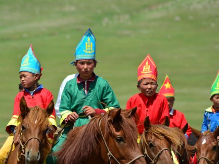 Eine Gruppe von Kindern in traditionellen bunten Gewändern reitet auf Pferden über eine grüne Wiese, voller Freude und Energie.