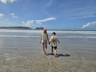 Zwei Kinder gehen nebeneinander am Strand entlang und halten sich an den Händen, während Wellen sanft an den Ufer schlagen.