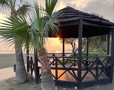 Ein Holzpavillon am Strand, umgeben von Palmen, während die Sonne hinter dem Horizont untergeht und den Himmel erleuchtet.