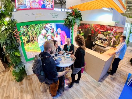 Eine Gruppe von Menschen diskutiert an einem Tisch in einem lebhaften Messestand, umgeben von tropischen Pflanzen.