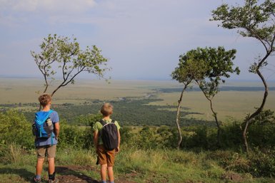 Zwei Kinder stehen auf einem Hügel und blicken auf die weite Landschaft der Masai Mara, umgeben von Bäumen.