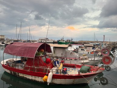 Ein rotes Fischerboot mit einem abgedeckten Steuerstand liegt ruhig im Hafen, umgeben von anderen Booten und einem bewölkten Himmel.