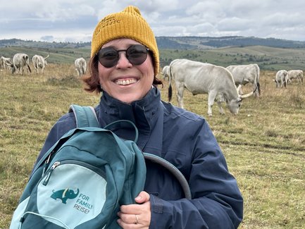 Eine lächelnde Frau mit einer gelben Mütze steht auf einer Wiese, umgeben von grasenden Kühen und einer hügeligen Landschaft.