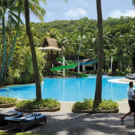 Umgeben von Palmen liegt ein glitzernder Pool, während ein Servicekraft eilends Handtücher bringt und das Resort belebt.