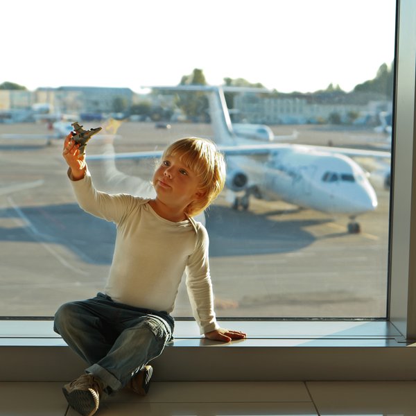 Ein kleiner Junge sitzt am Fenster eines Flughafens und hält ein Spielzeugflugzeug in der Hand, während er nach draußen schaut.