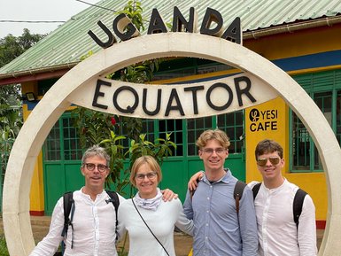 Eine Gruppe von vier Personen steht lächelnd am Äquator in Uganda, umgeben von bunten Gebäuden und Pflanzen.
