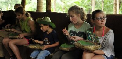Eine Gruppe von Kindern und Jugendlichen sitzt in einer Hütte und genießt traditionelle Speisen aus Blättern.