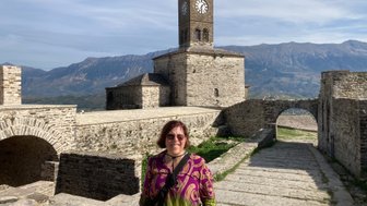 Eine Frau mit braunen Haaren steht lächelnd vor einer alten Steinburg mit einem Uhrturm und Bergen im Hintergrund.
