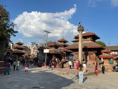 Ein belebter Platz in Kathmandu mit historischen Tempeln, Menschen, die sich versammeln, und einer beeindruckenden Wolkenformation.