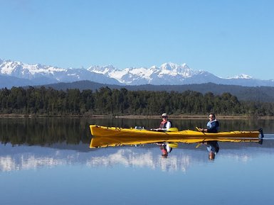 Zwei Personen paddeln in einem gelben Kajak auf einem ruhigen See, umgeben von majestätischen Bergen und klarem Himmel.