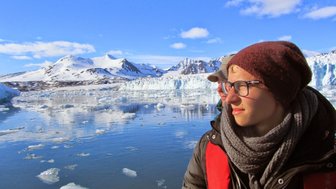 Eine Person mit Brille und Mütze blickt nachdenklich auf das ruhige Wasser, umgeben von schneebedeckten Bergen.