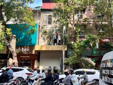 Eine belebte Straße in Hanoi, auf der viele Motorräder und Autos im Verkehr stehen, umgeben von grünen Bäumen.