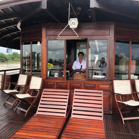 Ein Kapitän steht am Steuer eines Holzbootes, während Passagiere im Hintergrund die Aussicht genießen.