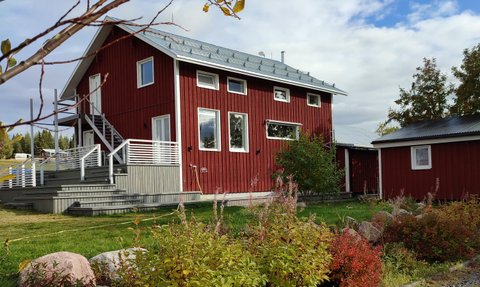 Ein rotes Holzhaus mit einem modernen Design steht in einer grünen Umgebung, umgeben von bunten Pflanzen und Steinen.