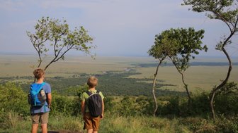 Zwei Kinder stehen auf einem Hügel und blicken auf die weite Landschaft der Masai Mara, umgeben von Bäumen.