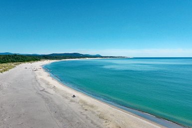Ein weitläufiger Strand mit feinem Sand erstreckt sich entlang des ruhigen, blauen Wassers unter einem strahlend blauen Himmel.