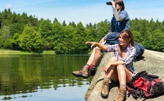 Zwei Jugendliche sitzen auf einem Felsen am Wasser, während einer mit einem Fernglas in die Ferne schaut.