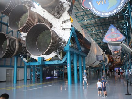 Im Kennedy Space Center stehen zwei Kinder fasziniert vor einem riesigen Raketenmotor, während Besucher im Hintergrund umhergehen.