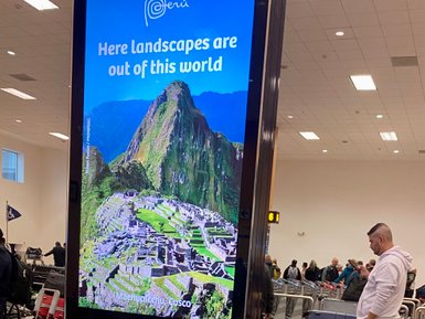 Ein Reisender steht am Gepäckband, während ein Werbeschild von Machu Picchu im Hintergrund leuchtet.