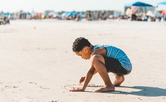 Ein Junge mit lockigem Haar sitzt am Strand und gräbt mit seinen Händen im Sand, während die Sonne scheint.