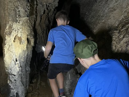 Zwei Kinder in blauen T-Shirts erkunden eine dunkle Höhle, während sie über große Steine klettern und eine Taschenlampe benutzen.