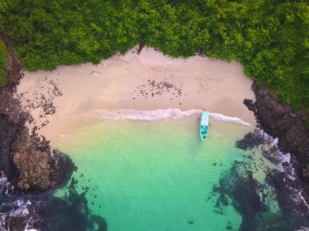 Ein malerischer Strand mit feinem Sand und klarem Wasser, umgeben von üppigem Grün und Felsen, bietet eine ruhige Atmosphäre.