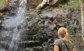 Eine Frau steht vor einem Wasserfall, der über eine felsige Klippe fließt, umgeben von üppigem Grün und Bäumen.