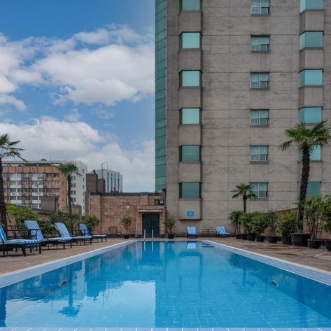 Ein klarer Pool spiegelt den Himmel wider, umgeben von modernen Gebäuden und Palmen, die eine entspannte Atmosphäre schaffen.