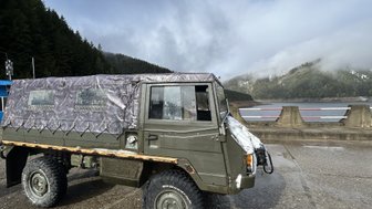 Ein geländegängiges Fahrzeug steht auf einer nassen Straße, umgeben von bewaldeten Hügeln und einem ruhigen See im Hintergrund.