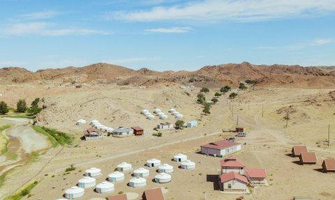 Eine weite Landschaft mit Jurten und kleinen Häusern, umgeben von sanften Hügeln und einem klaren blauen Himmel.